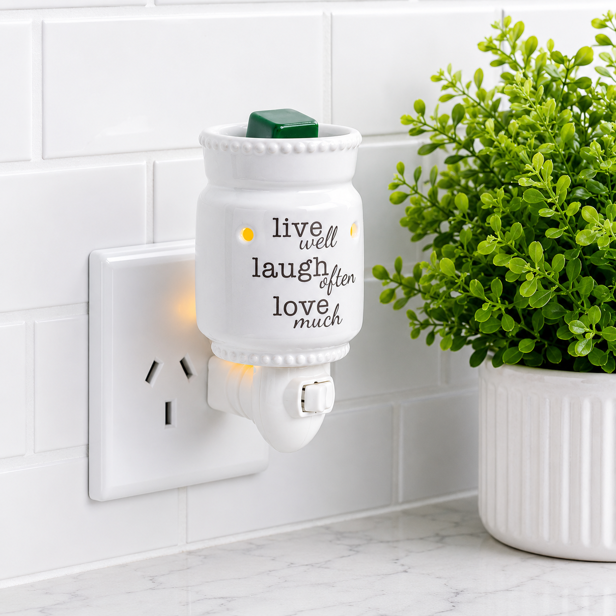 Wall-mounted diffuser with motivational quote next to a potted plant on a white tiled wall.