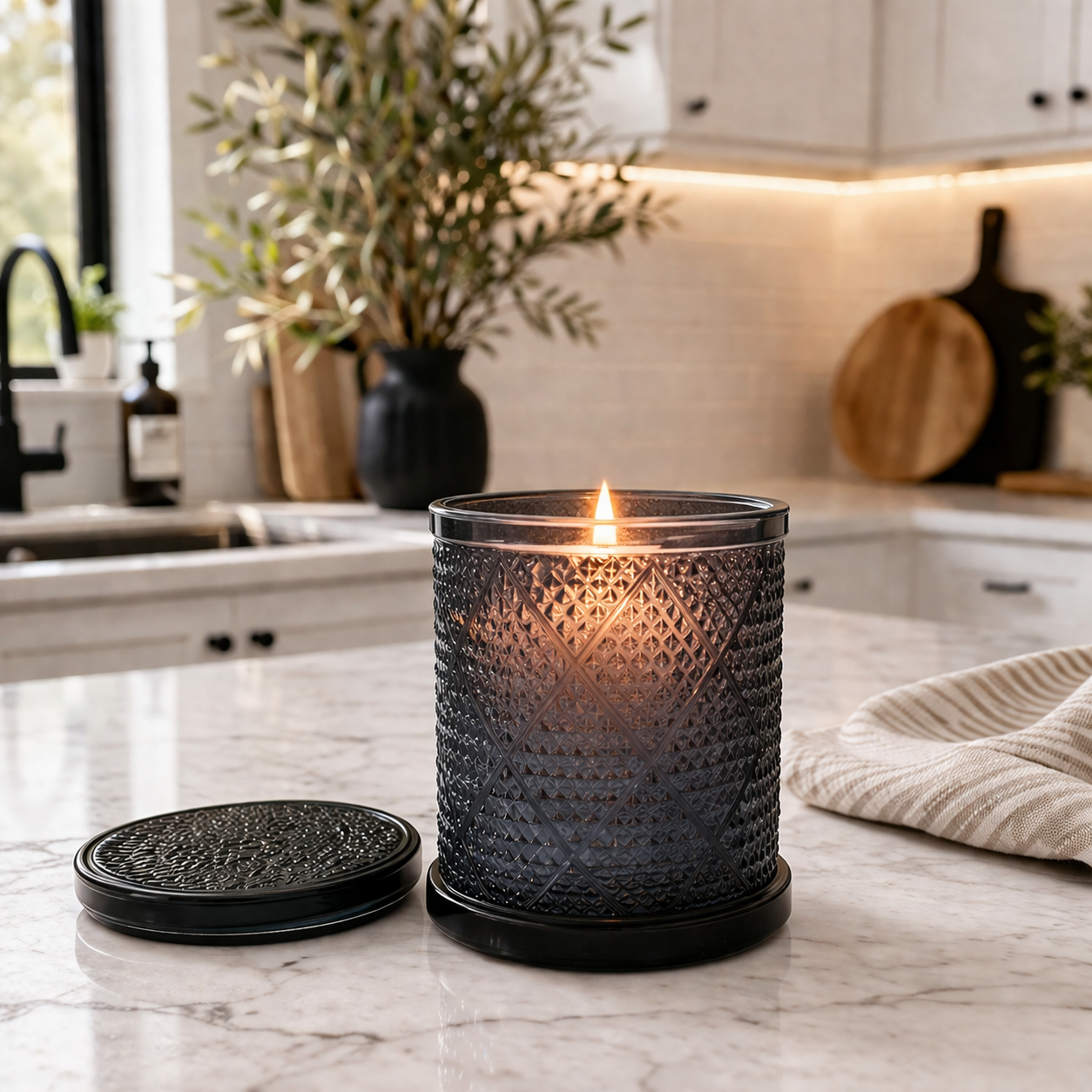 Decorative candle in a textured holder on a kitchen counter with a plant and kitchenware in the background.