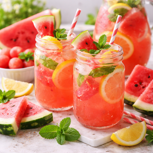 Two glasses of watermelon lemonade with slices of watermelon, lemon, and mint leaves on a marble surface.